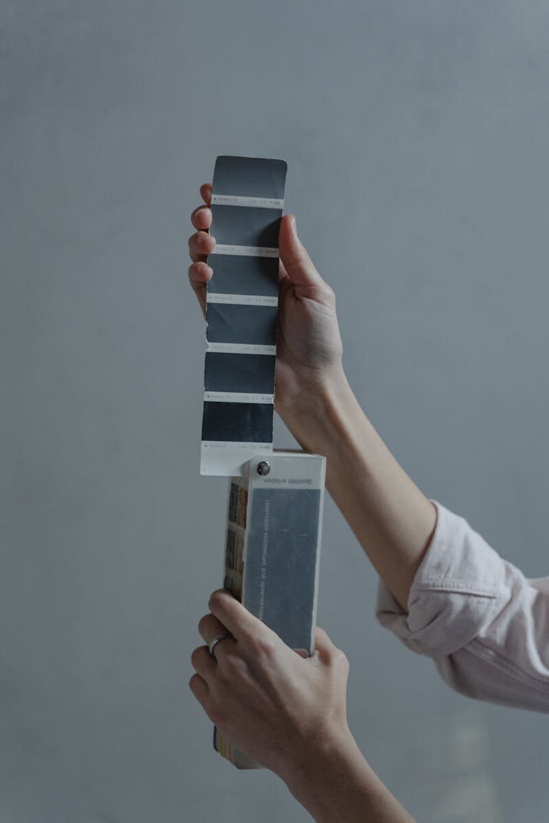 Close-up of hands holding and examining a gradient of paint color swatches against a gray background.